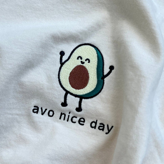 A white shirt with an embroidered avocado on the left chest with the quote "avo nice day".